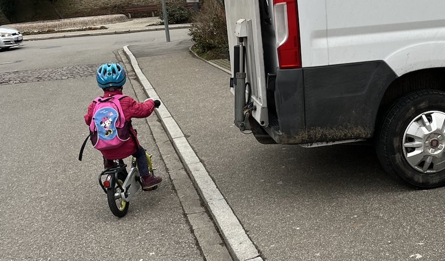 Ein Kind auf einem Laufrad muss auf die Straße ausweichen, weil ein verkehrswidrig geparkter Lieferwagen den Gehweg zustellt.