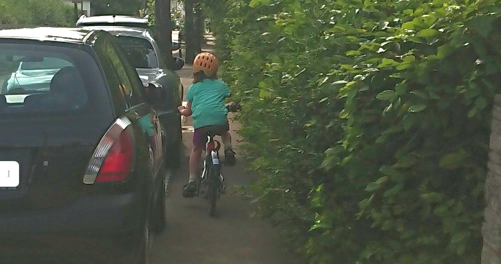 Ein kleines Kind fährt wie vorgeschrieben auf dem Gehweg. Es gibt keinen Platz: links parken Autos, rechts ragt eine ungeschnittene Hecke in den ohnehin zu knappen Platz.